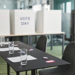 Sala preparada para votação, com mesas, cadeiras e cabines de privacidade organizadas para uma eleição.
