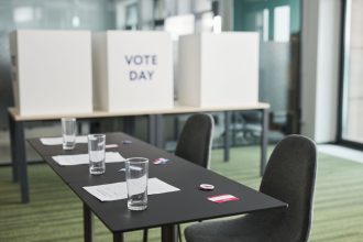 Sala preparada para votação, com mesas, cadeiras e cabines de privacidade organizadas para uma eleição.