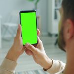 Homem segura um smartphone com a tela verde, usada como espaço para representar um aplicativo, dentro de casa.