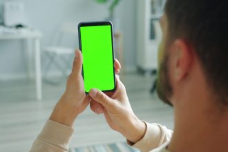 Homem segura um smartphone com a tela verde, usada como espaço para representar um aplicativo, dentro de casa.