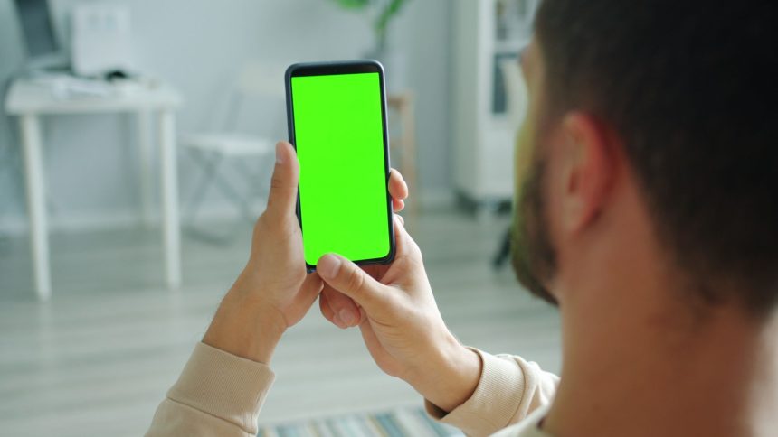 Homem segura um smartphone com a tela verde, usada como espaço para representar um aplicativo, dentro de casa.